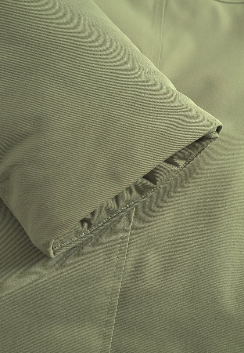 Tejido de chaqueta en verde oliva con una textura suave, que presenta un puño doblado y costuras bien hechas a lo largo de los bordes.