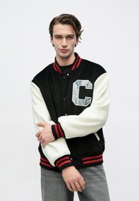 Νέος άνδρας που φορά ένα μαύρο και λευκό varsity μπουφάν με κόκκινες ρίγες στα μανίκια και ένα μεγάλο έμβλημα "C", στέκεται μπροστά σε απλό λευκό φόντο.