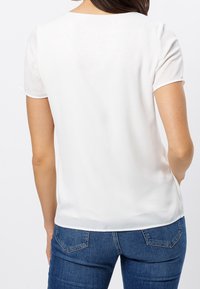 Weiße Bluse mit kurzen Ärmeln aus leichtem, glattem Stoff. Runde Ausschnitt und lockere Passform, kombiniert mit blauen Jeans.