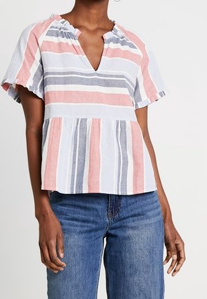 Kobieta w bluzce z krótkim rękawem z poziomymi paskami w kolorze różowym, szarym i białym, zestawiona z niebieskimi dżinsami na jednolitym tle.