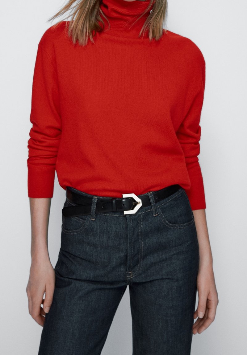 Pull à col roulé rouge en matériau doux, associé à un jean en denim sombre. Une ceinture noire avec une boucle en métal géométrique accentue la tenue.