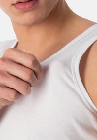 White cotton tank top with a ribbed texture and round neckline, showing a close-up of a hand gripping the fabric near the collar.