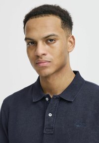 Tums zils polo krekls, izgatavots no teksturēta auduma, ar klasisku apkakli, trīs pogu aizdari un izšūtu logotipu uz krūšu daļas.