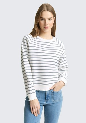 Femme aux longs cheveux bruns portant une chemise blanche à manches longues avec de fines rayures horizontales noires et un jean bleu, debout devant un fond uni.