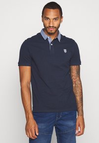 Marinblå polo med en chambray-krage och broderad logotyp. Korta ärmar och en skräddarsydd passform. Bärs med blå jeans, slät textur.