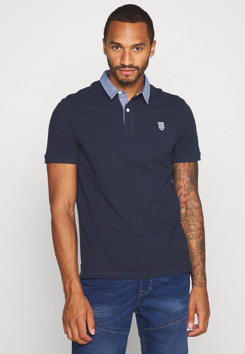 Marinblå polo med en chambray-krage och broderad logotyp. Korta ärmar och en skräddarsydd passform. Bärs med blå jeans, slät textur.