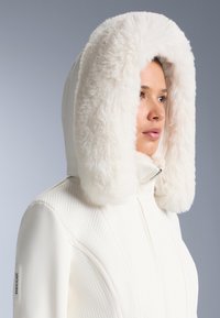 Îmbrăcăminte exterioară albă cu un corp texturat, având un glugă mare din blană artificială în crem deschis, închidere cu fermoar și logo RECCO® pe mânecă.