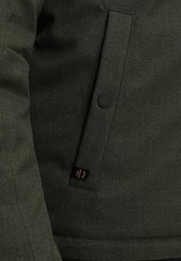 Dark green jacket made of textured fabric, featuring a buttoned flap pocket and a small brand label near the hem.