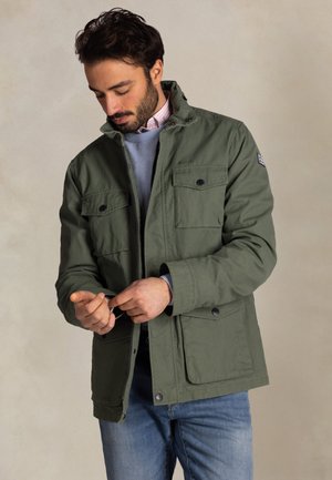 Man met een groene jas met klepzakken, een lichtblauwe trui en een spijkerbroek, die een mouwknop vastmaakt tegen een effen beige achtergrond.