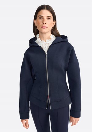 Felpa con zip di colore blu navy, dal design attillato, realizzata in tessuto morbido, con un ampio cappuccio e dettagli di cucitura sottili. Abbinata a una blusa in pizzo bianca sotto.