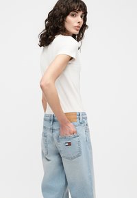 Γυναίκα με σγουρά μαλλιά, φοράει λευκό κοντομάνικο τοπ και ανοιχτό μπλε τζιν ψηλόμεσο με το χέρι στην πίσω τσέπη.