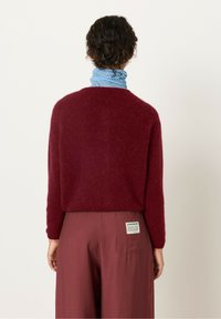 Burgundijska puhasta pulover s okroglo ovratnico, v kombinaciji z ohlapnimi maroon hlačami. Modro-bela črtasta ruta je ovita okoli vratu.