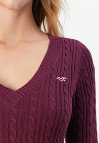 Žena na sebe má oblečený tmavo purpurový sveter s pleteným vzorom a výstrihom do V, na ľavej hrudi má malú bielu vtáčiu nášivku.