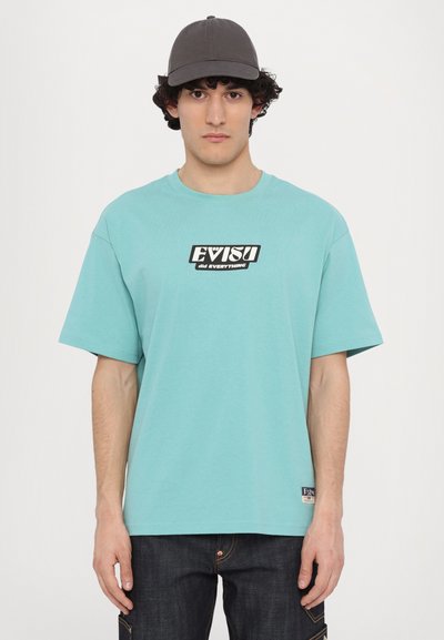 Τυρκουάζ κοντομάνικο T-shirt με μαύρο λογότυπο EVISU μπροστά, σε συνδυασμό με σκούρες τζιν παντελόνες και γκρι καπέλο μπέιζμπολ.