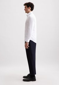 Weißes Langarmhemd mit Kragen, getragen mit marineblauen Hosen und schwarzen Schuhen. Hat einen slim fit und Knopfmanschetten. Minimalistisches Design.