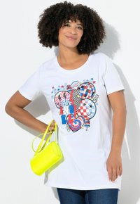 Hvid T-shirt med korte ærmer, der har et farverigt grafisk design med hjerter, stjerner og mønstre. Kombineret med en lille, lys gul crossbody taske.