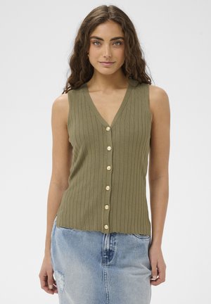 Ung kvinde med langt krøllet hår iført en olivengrøn ribstrikket ærmeløs bluse med knapper og en lyseblå denim nederdel.