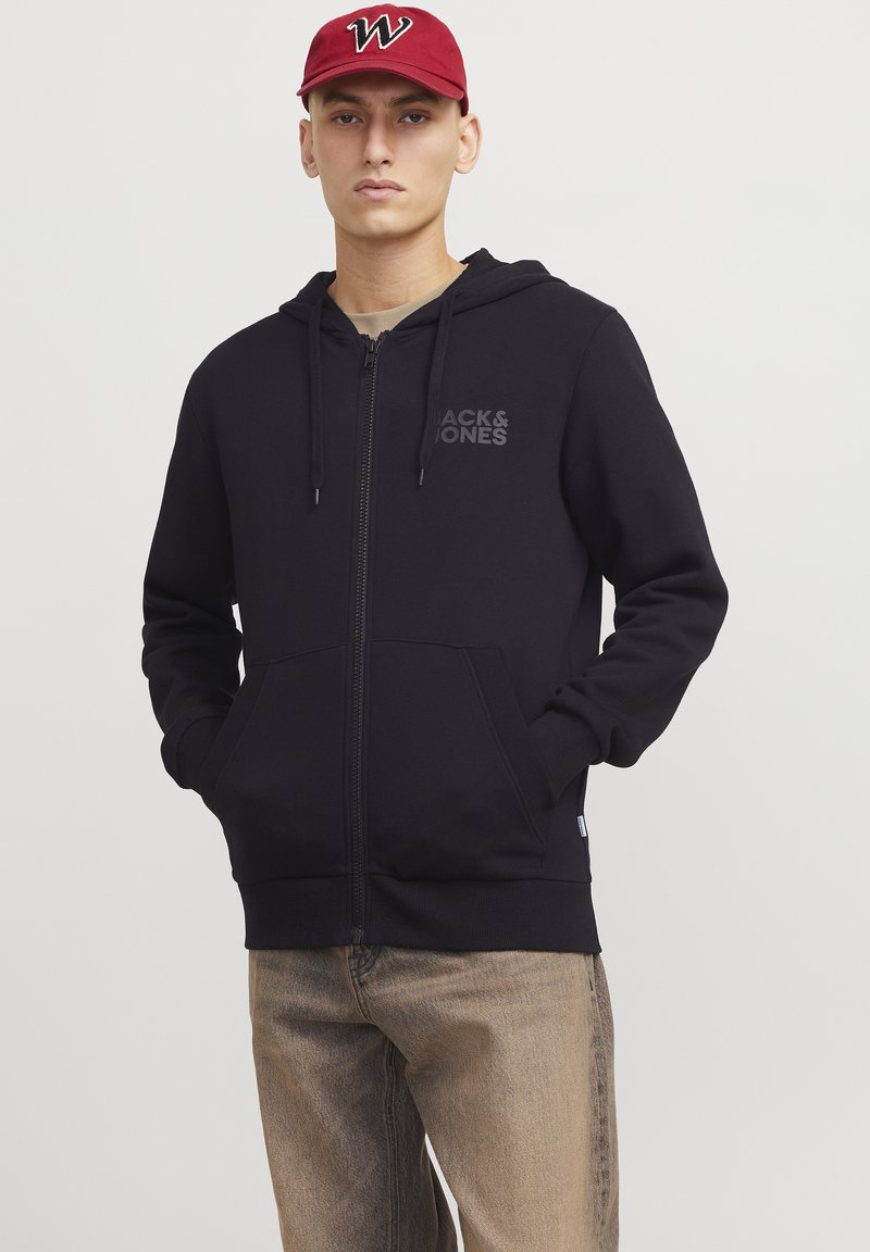 Sweat à zip noir avec une poche frontale, capuche à cordon, et un logo subtil sur la poitrine ; associé à un jean beige délavé.