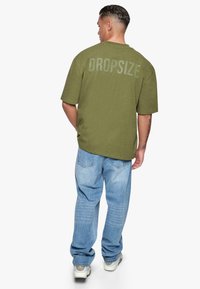 Olivengrünes, übergroßes T-Shirt mit dem Aufdruck "DROPSIZE" auf dem Rücken, kombiniert mit hellblauen, weiten Jeans und weißen Sneakers.