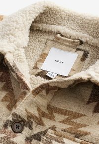 Chemise à col doublée de Sherpa en beige avec des motifs géométriques marrons ; elle possède une fermeture à boutons et une étiquette de marque à l'intérieur.