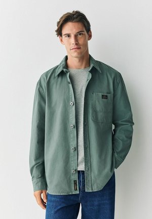 Hellgrünes Hemd mit Knöpfen und einem Cordkragen, Taschen auf der Brust und einem grauen T-Shirt darunter. Kombiniert mit blauen Jeans.