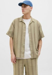 Chemise en toile à manches courtes beige avec des rayures texturées, devant à boutons, assortie à un short et à une casquette bleue. Tissu lisse et léger.
