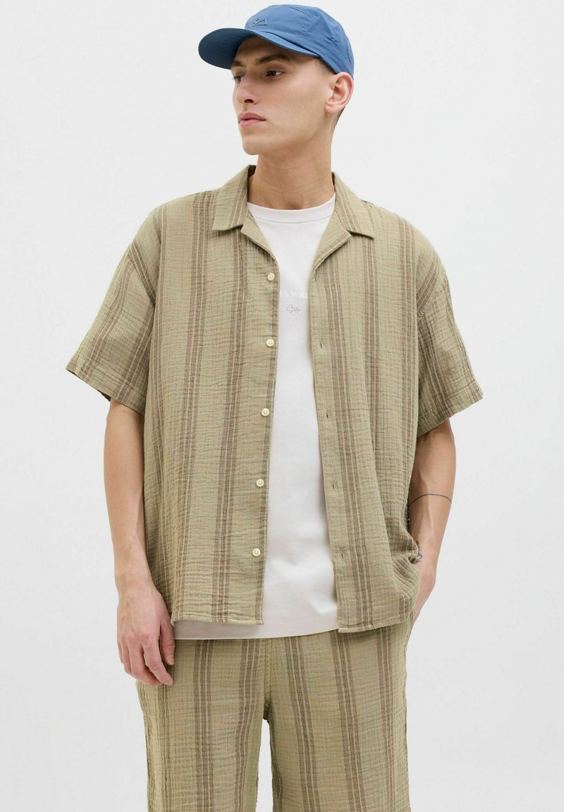 Chemise en toile à manches courtes beige avec des rayures texturées, devant à boutons, assortie à un short et à une casquette bleue. Tissu lisse et léger.