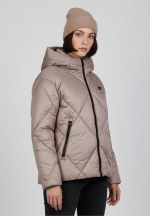 Giacca imbottita beige con un design a diamante trapuntato, cappuccio, chiusura a zip frontale e tasche laterali con accessori neri. Indossata con un berretto di maglia.