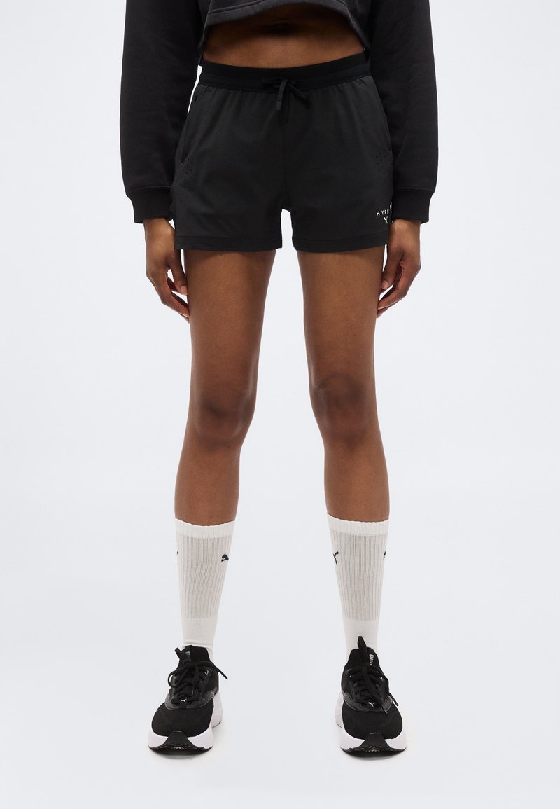 Άτομο που φοράει μαύρο αθλητικό σορτς, μαύρα αθλητικά παπούτσια, λευκές κάλτσες Puma που φτάνουν μέχρι την γάμπα και μαύρο κοντό φούτερ, στέκεται μπροστά σε ένα απλό φόντο.