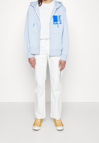 Felpa con cappuccio azzurro chiaro con cerniera frontale e tasca, maglietta bianca, pantaloni bianchi e scarpe da ginnastica gialle con suole bianche.