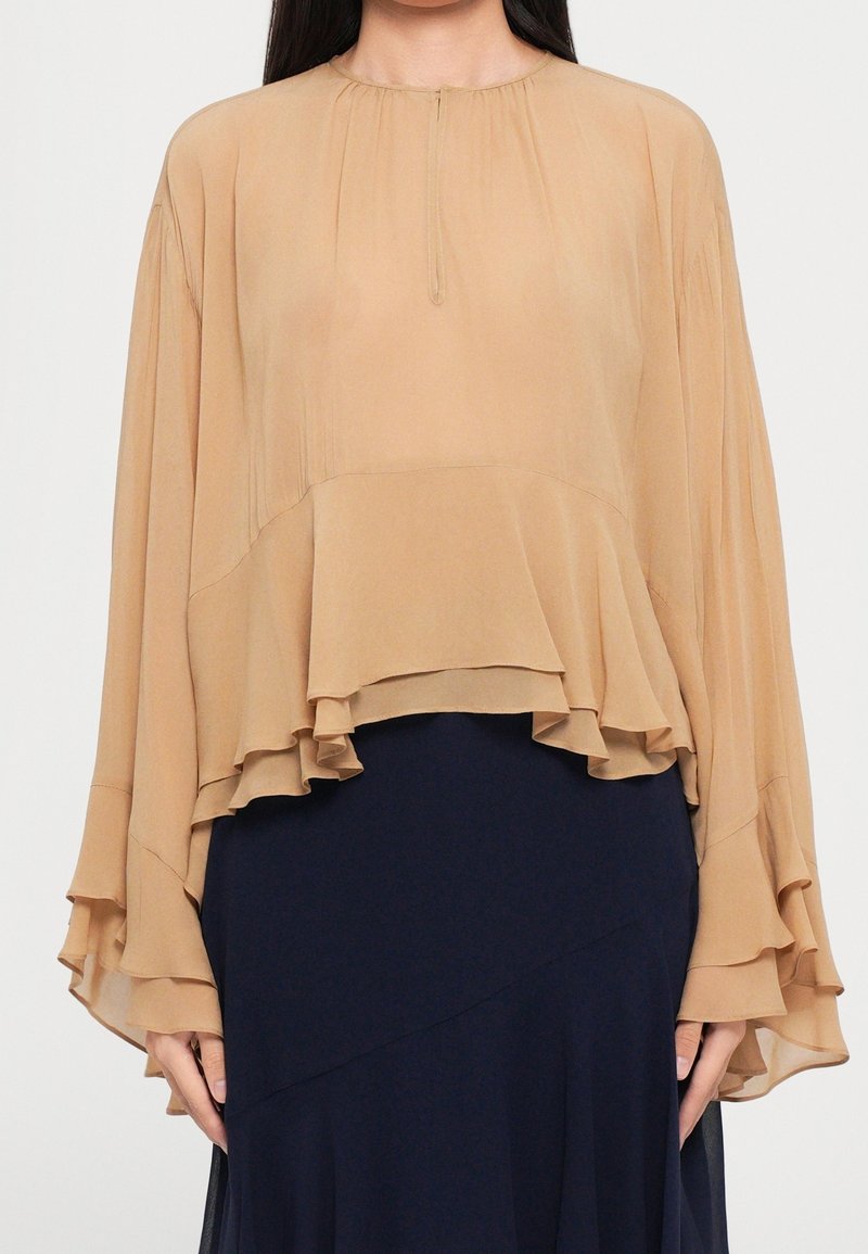 Femme portant un chemisier en mousseline ample, couleur beige, à manches longues et à volants superposés, sur une jupe bleu marine foncé, debout les bras le long du corps.