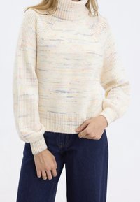 Pull à col roulé crème avec un tricot texturé parsemé de petites taches multicolores. Poignets et ourlet côtelés, associé à un jean bleu foncé.