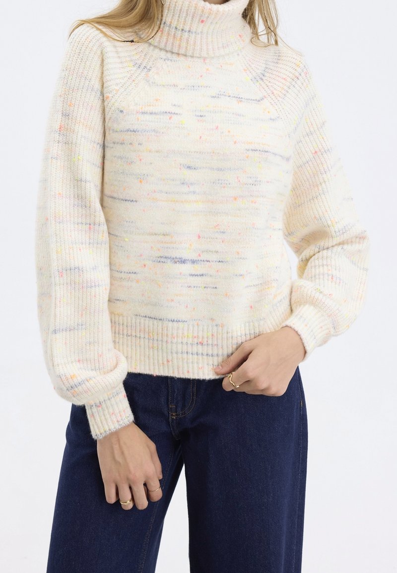 Pull à col roulé crème avec un tricot texturé parsemé de petites taches multicolores. Poignets et ourlet côtelés, associé à un jean bleu foncé.