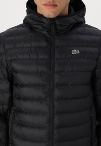 Černá prošívaná bunda s zapínáním na zip a kapucí. Má vodorovné prošívané části a malý zelený krokodýlí log na hrudi.