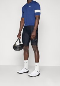 Maillot de cyclisme bleu pour homme à manches courtes, avec accent blanc, et short rembourré noir. Chaussures de cyclisme blanches et casque en main.