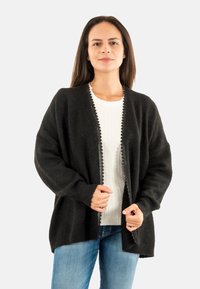 Cárdigan negro con una textura suave, de abertura frontal y bordes en forma de concha. Usado sobre un suéter blanco, combinado con jeans azules.