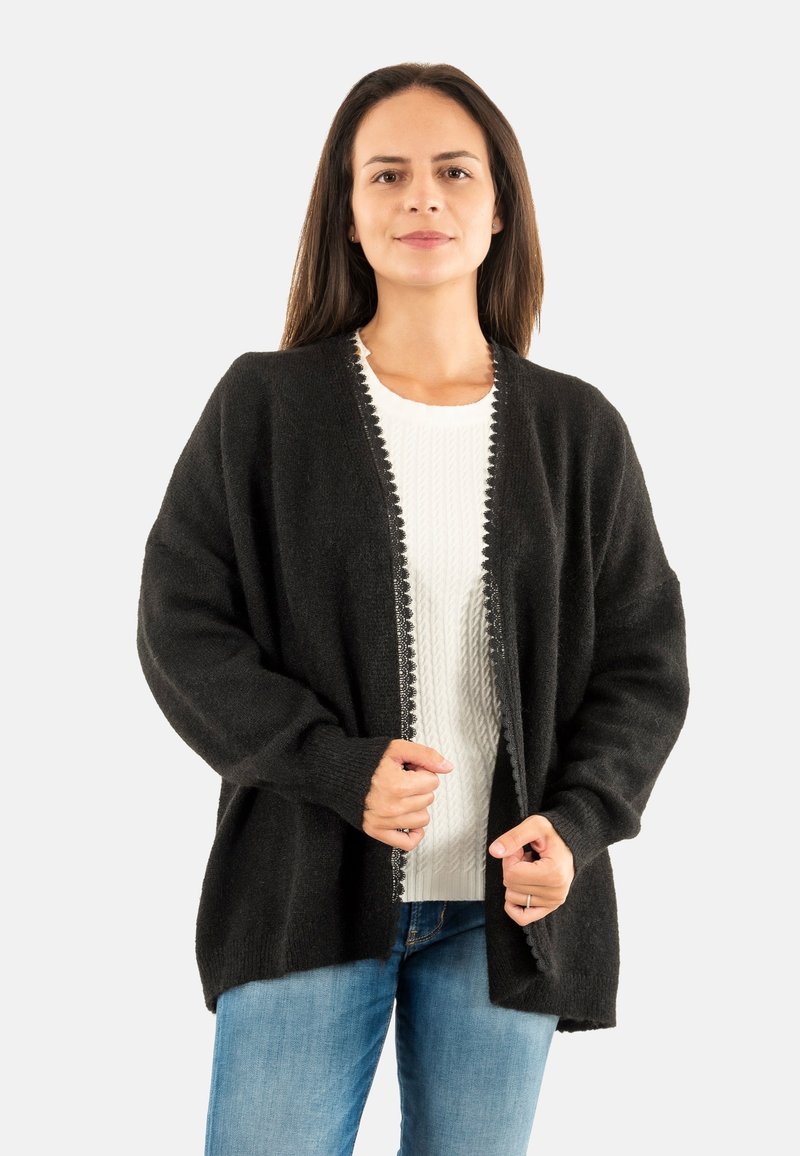 Cárdigan negro con una textura suave, de abertura frontal y bordes en forma de concha. Usado sobre un suéter blanco, combinado con jeans azules.