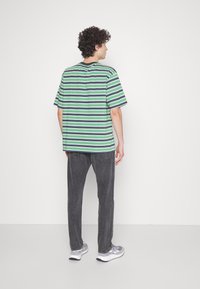 Gestreept t-shirt in groen, marineblauw en wit, met korte mouwen en een relaxte pasvorm. G gedragen met donkergrijze jeans en grijze sneakers.