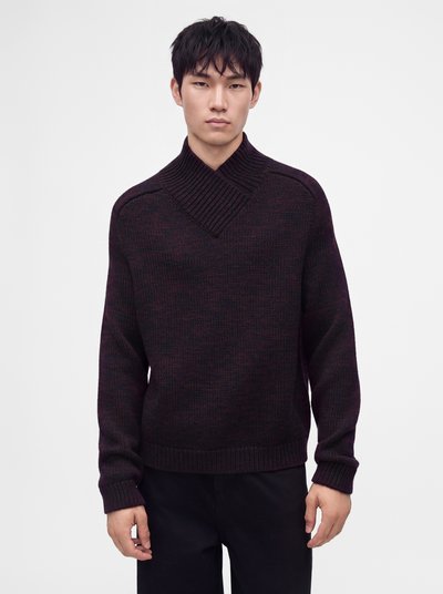 Homme aux cheveux courts et noirs portant un pull en maille foncée avec un large col châle côtelé et un pantalon noir, debout devant un fond uni.