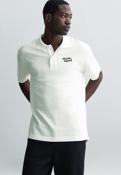 Polo bianco in tessuto a trama con colletto, caratterizzato da un logo ricamato scuro sul lato sinistro del petto. Indossato con pantaloni scuri.