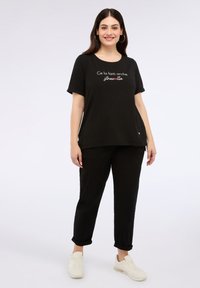 T-shirt nera a maniche corte con scritte bianche e dettagli rossi, abbinata a pantaloni neri. Modello che indossa sneakers bianche, in piedi su uno sfondo chiaro.