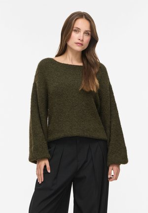 Donna con capelli castani lunghi, che indossa un maglione verde scuro largo e pantaloni neri plissettati, in piedi davanti a uno sfondo bianco semplice.