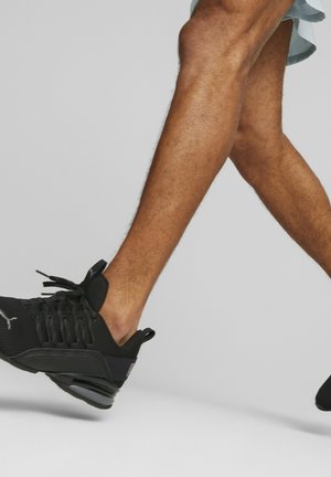 Benene af en person, der går, iført sorte Puma-sneakers og lyseblå shorts mod en ensfarvet lys grå baggrund.