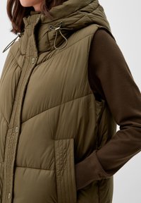 Olijfgroen gewatteerde vest met capuchon, ritssluiting en drukknopen, gequilt textuur en zijzakken, gelaagd over een bruine lange mouw shirt.