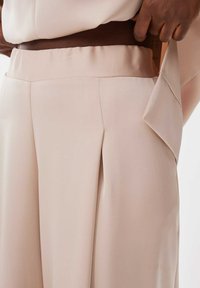 Beige op maat gemaakte broek van een gladde, lichte stof. Voorzien van een brede tailleband en voorplooien voor extra structuur en detail.