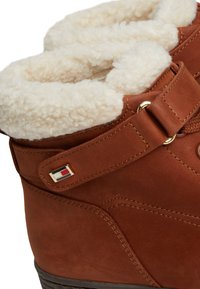 Stivali in suede marrone con un morbido rivestimento color crema, dotati di una cintura e accessori in oro. Un piccolo logo in rosso, bianco e blu è visibile sul lato.