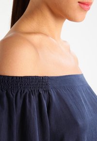 Navy off-the-shoulder top with smocked neckline and loose, flowing sleeves. Fabric appears lightweight with subtle texture.