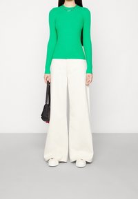 Top verde a maniche lunghe con coste abbinato a pantaloni bianchi a gamba larga. Tiene in mano una piccola borsa nera. Scarpe bianche con plateau. Sfondo semplice.