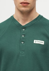 Άνδρας που φοράει πράσινο ανάγλυφο πουκάμισο henley με τρία κουμπιά και λευκή ετικέτα με το λογότυπο της Columbia στο στήθος.