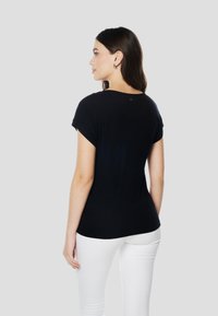 Fekete, rövid ujjú póló puha anyagból, kerek nyakkivágással és ívelt szegéllyel, fehér nadrággal viselve, hátulnézetben.